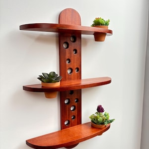 May include: A three-tiered, wooden wall shelf with a rich, reddish-brown finish. The shelf has a central support with circular cutouts and three curved shelves, each holding a small potted plant. The design is modern and suitable for displaying small items.