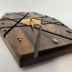May include: Geometric wooden wall clock with a dark brown finish and gold-colored hands and numerals. The clock face features a unique design with intersecting lines and a central wooden accent.