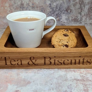 May include: A wooden tray with the words "Tea & Biscuits" engraved on the front. The tray has a white mug with a brown liquid inside and a chocolate chip cookie on it.