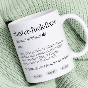 Könnte beinhalten: Eine weiße Keramiktasse mit einem schwarzen Design, auf dem "cluster-fuck-fixer" steht und der Begriff als ein Experte für Problemlösungen definiert wird, der jede Situation lösen kann, egal wie spektakulär sie in die Hölle und zurück geschraubt wurde. Die Tasse steht auf einer hellgrünen Strickdecke.