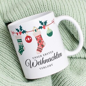 Könnte beinhalten: Weiße Keramik-Tasse mit einem Weihnachtsstrumpf-Design und dem Text "UNSER ERSTES Weihnachten VERLOBT". Die Tasse hat einen Henkel und ist mit Stechpalmen, Ornamenten und festlichen Strümpfen verziert. Die Tasse liegt auf einer grünen Strickdecke.