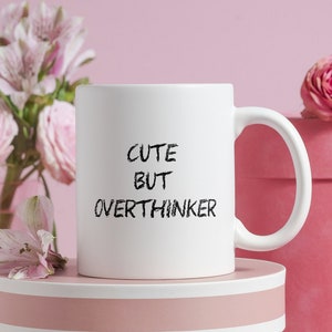 Könnte beinhalten: Weiße Keramiktasse mit schwarzem Text, der "Cute But Overthinker" auf einem rosa Hintergrund mit rosa Blumen und einer rosa Geschenkbox lautet.