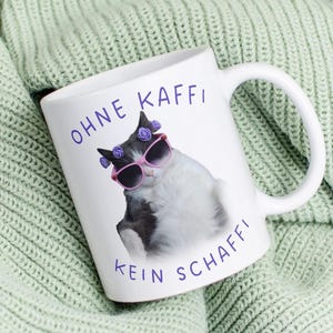 Pode incluir: Caneca de cerâmica branca com uma alça, apresentando um gato preto e branco usando óculos de sol rosa e detalhes de flores roxas. A caneca tem o texto "OHNE KAFFI KEIN SCHAFFI" em azul. A caneca está em um cobertor de malha verde.