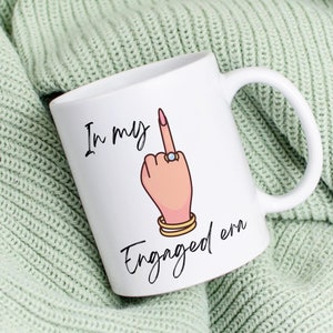 Könnte beinhalten: Weiße Keramiktasse mit einer Schwarzweißillustration einer Hand, die den Mittelfinger zeigt, mit einem Diamantring am Finger. Der Text "In my engaged era" ist unter der Illustration gedruckt.