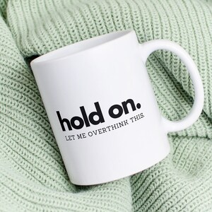 Könnte beinhalten: Weiße Keramiktasse mit schwarzem Text, der "hold on. let me overthink this" auf einer grünen Strickdecke lautet.