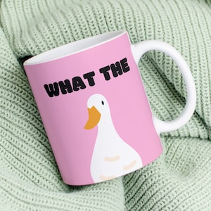 Könnte beinhalten: Eine rosa Keramiktasse mit einer weißen Gans und dem Text "WHAT THE" in schwarzen Buchstaben. Die Tasse steht auf einer grünen Strickdecke.