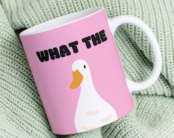 May include: A pink ceramic mug with a white goose and the text "WHAT THE" in black letters. The mug is sitting on a green knitted blanket.