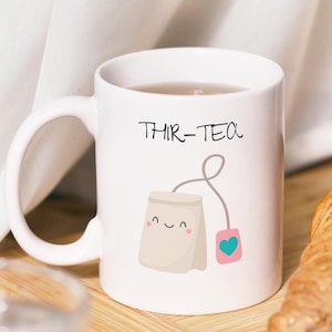 Könnte beinhalten: Eine weiße Keramiktasse mit einer Cartoon-Illustration eines Teebeutels und dem Text "Thir-Tea". Die Tasse ist mit Tee gefüllt und steht auf einer Holzoberfläche neben einem Croissant.