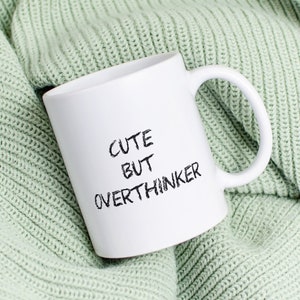 Könnte beinhalten: Weiße Keramiktasse mit dem Text "Cute but Overthinker" in schwarzen Buchstaben, die auf einer hellgrünen Strickdecke liegt.