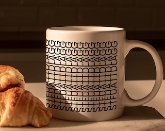 Può includere: Una tazza di ceramica bianca con un motivo geometrico nero e la parola "BLA" in verticale. Due croissant dorati riposano su un quadrato di marmo bianco. La tazza è sul marmo, con uno sfondo di cucina.
