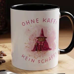 Könnte beinhalten: Weißer Keramikbecher mit schwarzem Innenraum und Henkel. Der Becher zeigt eine skurrile Illustration einer Katze im Zauberkostüm mit dem deutschen Text "OHNE KAFFI KEIN SCHAFFI" in Rosa.