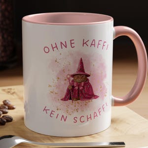 Könnte beinhalten: Weißer Keramikbecher mit rosa Innenraum und Henkel. Der Becher zeigt den Text "OHNE KAFFI KEIN SCHAFFI" in Rosa, zusammen mit einer Illustration einer Katze in einem Zaubererkostüm. Ein Löffel und ein Macaron liegen auf der Holzoberfläche.