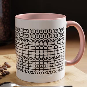 May include: White ceramic mug with a pink interior and handle. The mug features a black geometric pattern around the exterior. A spoon and a macaron are on a wooden surface next to the mug.