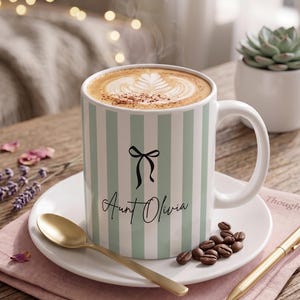 Pode incluir: Uma caneca de cerâmica branca com riscas verticais verde-claras, com um gráfico de laço preto e o texto "Aunt Olivia" em letra cursiva. A caneca está cheia de café com latte art e assenta num pires branco com uma colher dourada.