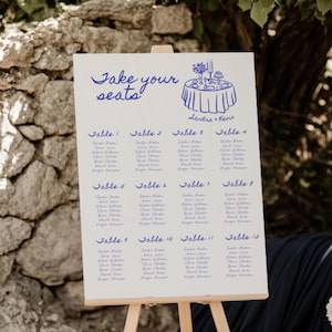 May include: A white seating plan on a wooden easel, featuring blue text that says "Take your seats" and table assignments. A floral illustration is above the names. The scene is outdoors with flowers and a stone wall.