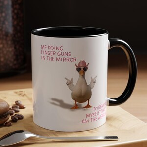 Könnte beinhalten: Weiße Keramik-Tasse mit schwarzem Innenraum und Griff. Die Tasse zeigt eine Cartoon-Ente mit Sonnenbrille und Cowboyhut, mit dem Text "ME DOING FINGER GUNS IN THE MIRROR" und "TO REMIND MYSELF THAT I AM THE WEAPON."
