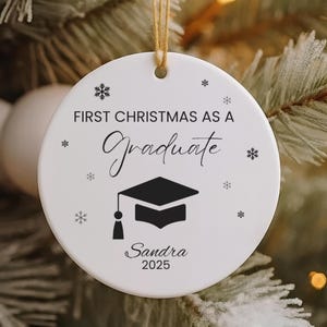Könnte beinhalten: Weißer Keramik-Weihnachtsschmuck mit dem Text "FIRST CHRISTMAS AS A Graduate" und einer Grafik einer Abschlusskappe. Der Schmuck enthält den Namen "Sandra" und das Jahr "2025". Er hängt an einem Weihnachtsbaum.