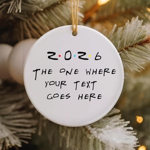 Op de afbeelding: Een rond wit keramieken ornament dat aan een beige touwtje hangt. Het ornament bevat de tekst 2026 met gekleurde stippen tussen de cijfers en de zwarte tekst THE ONE WHERE YOUR TEXT GOES HERE in een handgeschreven lettertype op het oppervlak.