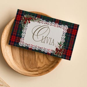 Könnte beinhalten: Eine rechteckige Karte mit einem rot-grün-blauen Schottenmuster und einem weißen Spitzenrand. In der Mitte steht der Name Olivia in goldener Schreibschrift, akzentuiert durch Illustrationen von Stechpalmen, Tannenzapfen und roten Schleifen.