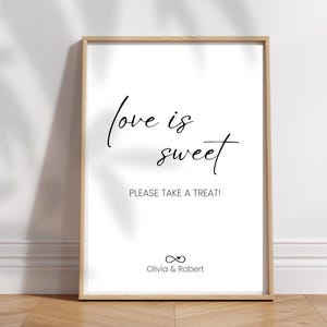 Könnte beinhalten: Ein gerahmtes Schild mit den Worten "love is sweet" in eleganter Schrift, zusammen mit "PLEASE TAKE A TREAT!" und den Namen "Olivia & Robert". Das Schild hat einen hellen Holzrahmen und ist vor einem weißen Hintergrund platziert.