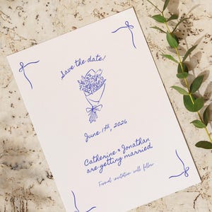 Könnte beinhalten: Eine weiße Save-the-Date-Karte mit blauer Schrift und einer Illustration eines Blumenstraußes. Der Text lautet "Save the date!" und "June 17th, 2026." Auf der Karte steht auch "Catherine & Jonathan heiraten."