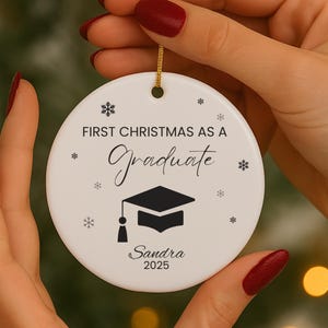 Könnte beinhalten: Weißer Keramik-Weihnachtsschmuck mit dem Text "FIRST CHRISTMAS AS A Graduate" und einer Grafik einer Abschlusskappe. Der Schmuck enthält auch den Namen "Sandra" und das Jahr "2025".
