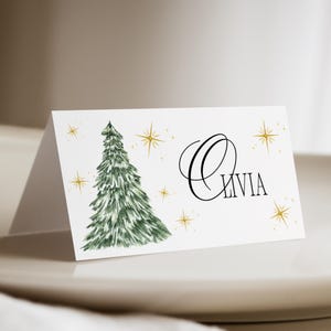 Könnte beinhalten: Eine weiße Tischkarte mit einer Aquarellillustration eines grünen Weihnachtsbaums und goldenen Sternen. Der Name "Olivia" ist in schwarzer Schrift geschrieben. Ideal für festliche Tischdekorationen.