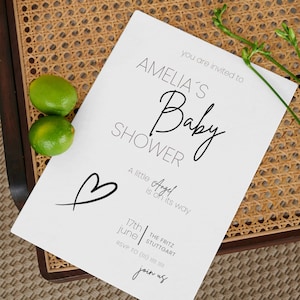 Könnte beinhalten: Eine weiße Babyparty-Einladung mit dem Text "AMELIA'S Baby SHOWER" in eleganter Schrift. Die Einladung enthält den Satz "A little Angel is on its way" und ein Herzsymbol. Zwei grüne Limetten und Kräuterzweige sind ebenfalls zu sehen.