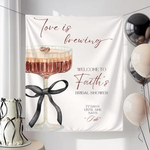 Könnte beinhalten: Ein weißes Banner mit dem Text "Love is brewing" und "Welcome to Faith's Bridal Shower". Eine Aquarellillustration eines Cocktailglases mit einer schwarzen Schleife ist zu sehen. Ballons und ein Kuchen sind ebenfalls sichtbar.