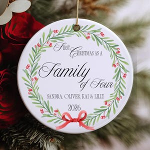 Könnte beinhalten: Ein rundes, weißes Keramikornament mit einem grünen Kranz aus Blättern und roten Beeren sowie einer roten Schleife unten. Der Text lautet: First Christmas as a Family of Four, Sandra, Oliver, Kai & Lilly, und 2026. Es hängt an einer goldenen Schnur.