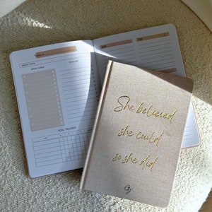 Könnte beinhalten: Ein beigefarbenes Leinen-Notizbuch mit goldener Aufschrift "She believed she could so she did" und der Signatur "Cy". Das Notizbuch ist auf einer Seite mit einem Wochenplaner und einem Ziel-Tracker geöffnet.