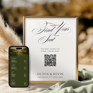 以下が含まれることがあります： 「Find Your Seat」と書かれたエレガントな筆記体の金色の額縁のサイン、QRコード、結婚式の詳細。スマートフォンには座席表が表示されています。背景はぼやけており、柔らかい緑色の布があります。