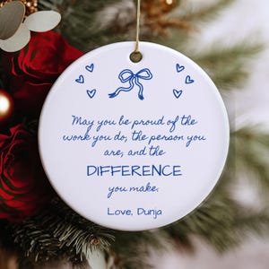 Könnte beinhalten: Ein rundes, weißes Keramikornament mit einer blauen Strichzeichnung einer Schleife und sechs kleinen Herzen. Der Text lautet: May you be proud of the work you do, the person you are, and the DIFFERENCE you make. Love, Dunja. Mit goldener Kordel.