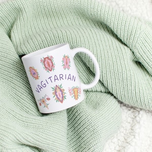 May include: White ceramic mug with a pink, purple, and orange floral design and the word "Vagitarian" in purple lettering.