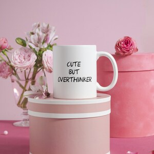 May include: White ceramic mug with black text that reads "Cute but Overthinker" on a pink background with pink flowers and a pink gift box.