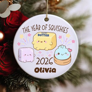 Könnte beinhalten: Ein rundes, weißes Keramikornament mit dem Text THE YEAR OF SQUISHIES, 2026 und dem Namen Olivia. Das Design zeigt Illustrationen eines gelben Stücks gesalzener Butter, eines hellvioletten Würfels und eines hellblauen Dumplings, alle mit Gesichtern.