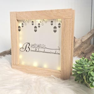 Könnte beinhalten: Ein hellbrauner Holzrahmen mit einer LED-Leuchtbox. Der weiße Hintergrund zeigt den arabischen Text "Bismillahir Rahmanir Rahim" sowie dekorative Laternen und Sterne. Der Rahmen besteht aus hellem Holz.
