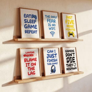 May include: Six framed prints with gaming quotes and graphics on wooden shelves. The prints feature phrases like "Eat Sleep Game Repeat," "The Only Fear Is No Wifi," and "Gamers Don't Die They Respawn." The frames have a wooden finish.