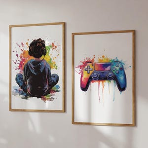 May include: Two framed watercolour prints. One shows a person wearing headphones, seated with their back to the viewer, surrounded by colourful paint splatters. The other depicts a vibrant, rainbow-coloured video game controller with paint drips.