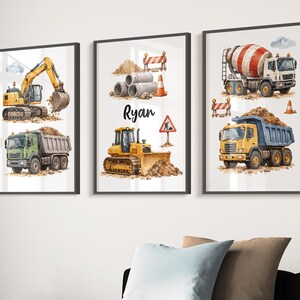 May include: Three framed wall art prints featuring watercolour illustrations of construction vehicles including a yellow excavator, a green dump truck, a yellow bulldozer, and a cement mixer. The centre print includes the name Ryan in black text.