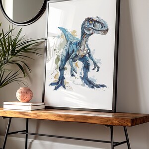 Puede incluir: Un cuadro enmarcado con una ilustraci&oacute;n de acuarela de un dinosaurio azul. La obra de arte tiene un fondo blanco con salpicaduras de pintura azul y verde. El cuadro con marco negro se exhibe sobre un banco de madera.