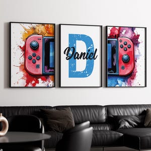 May include: Three framed prints with a watercolour background. The left and right prints feature a red and black video game controller. The centre print features a blue letter D with the name Daniel in white text.