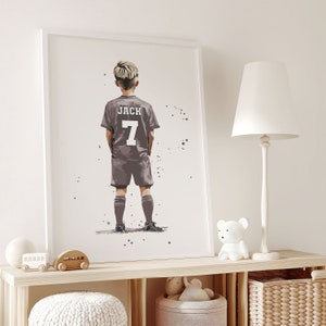 K&ouml;nnte beinhalten: Ein wei&szlig; gerahmter Druck eines jungen Jungen, der ein graues Fu&szlig;balltrikot mit dem Namen "JACK" und der Nummer "7" auf dem R&uuml;cken tr&auml;gt. Der Junge steht mit dem R&uuml;cken zum Betrachter und blickt nach links. Der Hintergrund ist wei&szlig; mit einigen grauen Punkten.