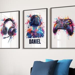 May include: Three framed art prints with a gaming theme. One print features a headset, another a person wearing a headset with the name "DANIEL", and the third a game controller. Each image has a colourful splatter paint effect.