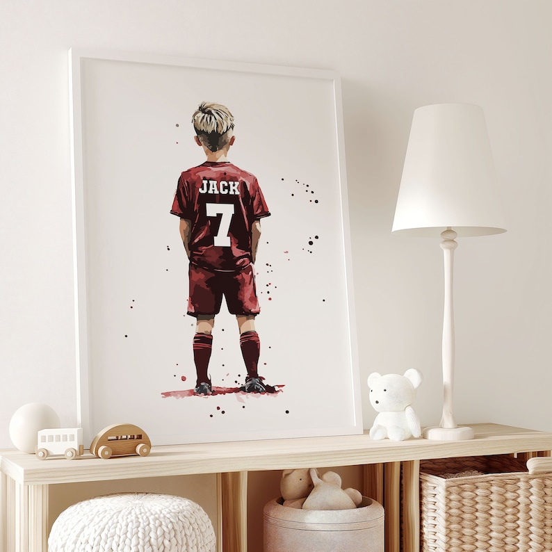 K&ouml;nnte beinhalten: Ein wei&szlig; gerahmter Druck eines jungen Jungen, der ein rotes Fu&szlig;balltrikot mit dem Namen "JACK" und der Nummer "7" auf dem R&uuml;cken tr&auml;gt. Der Junge steht mit den H&auml;nden in den Taschen und blickt von der Kamera weg. Der Hintergrund ist ein wei&szlig;er Aquarell-Wasch mit schwarzen Spritzern.