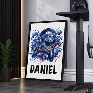 May include: A framed print with a gaming theme, showcasing a controller and headphones in shades of blue and purple. The name "DANIEL" is written in bold black letters. The artwork is set against a white background, with a black frame.