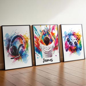May include: Three framed wall art prints featuring vibrant watercolour splashes. The left print shows gaming headphones, the centre print depicts a person holding a game controller with the name James below, and the right print displays a game controller.
