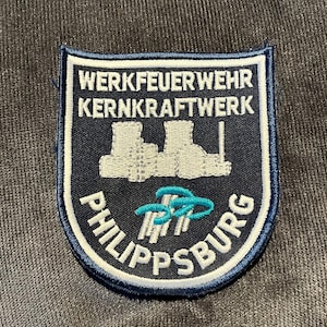 May include: A navy blue and white patch with the text "WERKFEUERWEHR KERNKRAFTWERK PHILIPPSBURG". The patch features a stylized illustration of a power plant in white and teal accents. The patch is rectangular with rounded corners.