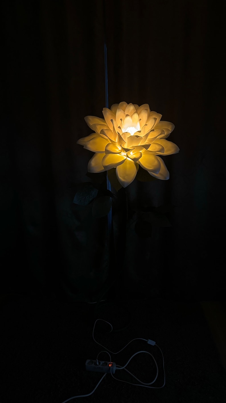 Puede incluir: Una l&aacute;mpara de papel con forma de flor blanca con una bombilla de luz blanca c&aacute;lida. La l&aacute;mpara est&aacute; sobre un soporte negro y tiene un cable blanco.