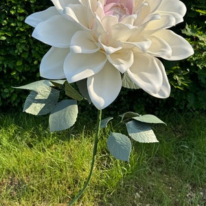 Puede incluir: Una gran flor de dalia artificial blanca con un centro rosa, en un tallo verde con hojas, de pie sobre una base verde en un &aacute;rea cubierta de hierba.